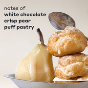 COCOA BLANCO fragrance notes displayed with a white chocolate-drizzled pear and two powdered puff pastries on a silver tray, with a spoon behind them and black text that reads 