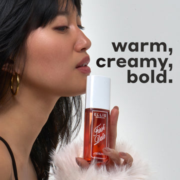 Woman with black hair and bangs wearing gold hoop earrings, a black tank top, and fuzzy, white sweater holds FIRST DATE Perfume Mist 90ml against her chin with black text that reads 