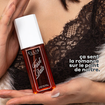 Un mannequin aux cheveux noirs, portant un haut en dentelle noire et un pull blanc duveteux, tient le parfum FIRST DATE Perfume Mist 90ml entre ses deux mains devant sa poitrine, avec un texte blanc indiquant « ça sent la romance sur le point de nattre ».