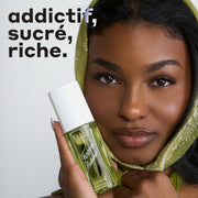 Femme aux cheveux noirs portant un foulard vert sur la tête et tenant une bouteille de brume parfumée FOREVER PISTACHIO 90 ml contre sa joue sur un fond blanc avec un texte en noir et blanc qui dit « addictif, sucré, riche. »