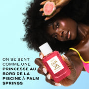 Une femme aux cheveux bruns portant un haut de bikini rose et jaune tient un GUAVA GRANITA Eau de Parfum 50ml couvert de gouttes d'eau devant elle dans une main et une tranche de goyave dans l'autre main près de sa tête, sur un fond bleu avec un texte noir qui dit « On se sent comme une princesse au bord de la piscine à Palm Springs. »