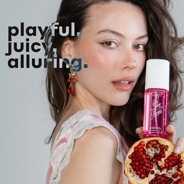Model with brown hair wearing pink silk and white lace tank top and pink earrings holds LOVE POM Perfume Mist 90ml and half of a pomegranate against a gray background with black and gray text that reads 