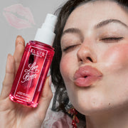 Woman with brown hair wearing pink silk and white lace tank top and pink earrings making a kissing face and holding a bottle of LOVE POM Perfume Mist 90ml without cap against a gray background with a faint pink lipstick outline