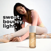 Model with brown hair wearing a pink top, yellow leotard, white tights, and pink ballet slippers sits and stretches forward with a MARSHMALLOWS Perfume Mist 240ml sitting on the white floor in front of her on a white background with white, black, and blue text that reads 