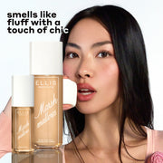 Model with brown hair wearing a peach top with a pink rose eats a marshmallow with MARSHMALLOWS Perfume Mist 240ml and 90ml in front of her on a white background with black text that reads 