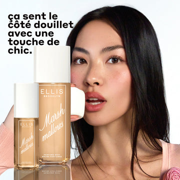 Modèle aux cheveux bruns portant un haut pêche avec une rose rose mange une guimauve avec le parfum en brume MARSHMALLOWS 240 ml et 90 ml devant elle sur un fond blanc avec un texte noir qui dit « ca sent le cote douillet avec une touche de chic »