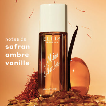 Vaporisateur de parfum Miss Amber 90 mL avec un bouchon blanc, entouré de liquide ambre, de filaments de safran, de gousse de vanille et de cailloux sur un fond pêche chaud. Le texte à gauche indique « notes de safran, ambre, vanille ».