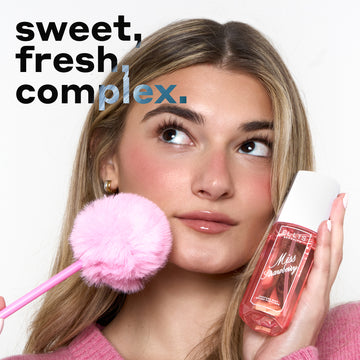 Woman with blonde hair in a pink sweater and gold hoop earrings looks up and holds MISS STRAWBERRY Perfume Mist 90ml against one cheek and a pink pen with a pink pompom on the end against the other cheek with black and blue text that reads 