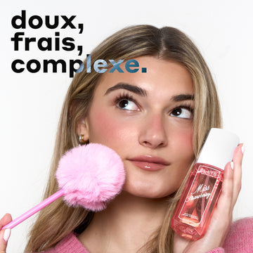 Femme aux cheveux blonds portant un pull rose et des boucles d'oreilles créoles dorées, regardant vers le haut et tenant le parfum MISS STRAWBERRY Perfume Mist 90ml contre une joue et un stylo rose avec un pompon rose au bout contre l'autre joue, avec un texte noir et bleu qui dit « doux, frais, complexe. » le tout sur un fond blanc.