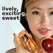 Model with black hair wearing orange tank top and headband holds NEW CRUSH Perfume Mist 90ml and slice of blood orange against white background with black and white text that reads 