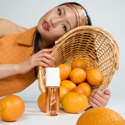 Woman with black hair wearing an orange and blue headband and tank top leaning against a basket filled with oranges behind a bottle of NEW CRUSH Perfume Mist 90ml and oranges against white background