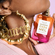 Gros plan d'une femme portant un pull rose et des boucles d'oreilles créoles en or où est écrit « Queen », tenant le parfum QUEENS CARAMEL Eau de Parfum 50 ml sans le bouchon sous son menton sur un fond rose
