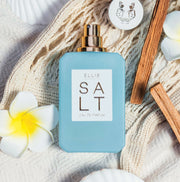 SALT Eau de Parfum 50ml sans bouchon posé sur une couverture blanche en tissu tissé à côté de fleurs blanches et jaunes, bouchon avec un motif léopard argenté, et des bâtons de bois