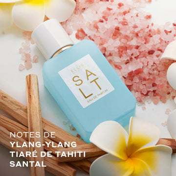 SALT Eau de Parfum 50ml est posé à côté de fleurs blanches et jaunes, de sel rose et de bâtons de bois sur fond blanc avec du texte blanc qui dit « Notes de ylang-ylang, tiare de tahiti, santal »