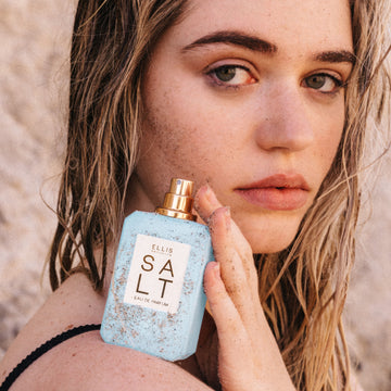 Femme aux cheveux bruns et mouillés portant un bikini noir tient le parfum SALT Eau de Parfum 50 ml sans le bouchon sur son épaule, le tout couvert de sable, sur fond de plage bronzée