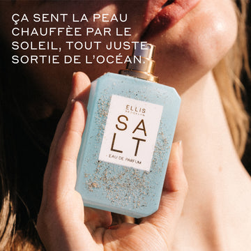 Femme aux cheveux blonds tenant SALT Eau de Parfum 50 ml sans bouchon, couvert de sable, contre son menton, baignée de soleil avec un texte blanc qui dit « Ça sent la peau chauffée par le soleil, tout juste sortie de l’océan. »