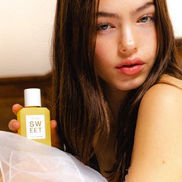 Une femme aux cheveux bruns portant une jupe en gaze blanche tient le parfum SWEET Eau de Parfum 50ml devant un fond marron et blanc