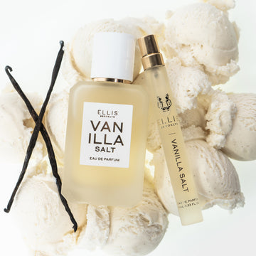 Gousses de vanille, sel de vanille 50 ml Eau de Parfum et sel de vanille 10 ml Eau de Parfum en vaporisateur de voyage reposant sur des boules de glace à la vanille, sur fond blanc.