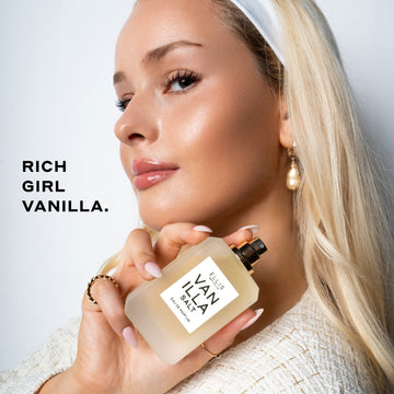 Model with blond hair and nude nail polish wearing white headband, pearl earring, gold rings, and cream boucle shirt holding Vanilla Salt 50ml Eau De Parfum without cap near shoulder on white background with black text that reads 