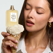 A woman with black hair wearing a white shirt and gold hoop earrings holds a vanilla ice cream cone with VANILLA SALT Eau de Parfum 50ml without cap on top all on a gray background.