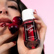 Model with brown hair and red nails looks at the camera while holding VANILLA SIN 90ml perfume mist and 2 cherries against her cheek on a pink background