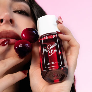Model with brown hair and red nails looks at the camera while holding VANILLA SIN 90ml perfume mist and 2 cherries against her cheek on a pink background