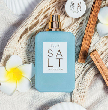 SALT Eau de Parfum 50ml without cap lays on white woven blanket next to white and yellow flowers, cap with silver leopard design, and sticks of wood