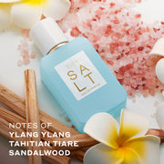 SALT Eau de Parfum 50ml lays next to white and yellow flowers, pink salt, and sticks of wood against white background with white text that reads 