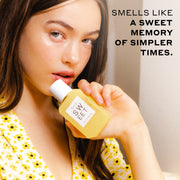 Woman with brown hair wearing a white top with yellow flowers holds SWEET Eau de Parfum 50ml against her chin against an off-white background with black text that reads 