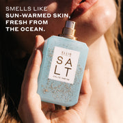 Woman with blonde hair holds SALT Eau de Parfum 50ml without cap covered in sand against her chin bathed in sunlight with white text that reads 