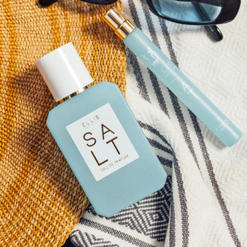 SALT Eau de Parfum 50ml and 10ml lay on white and gray and yellow towels next to black sunglasses with blue lenses