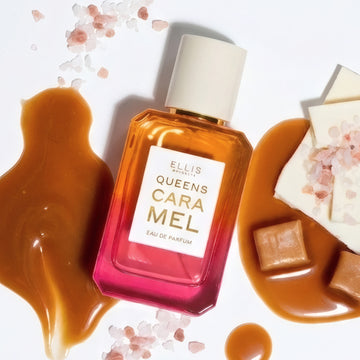 QUEENS CARAMEL Eau de Parfum 50ml est posé à côté de trois flaques de caramel, deux tas de sel rose, deux bonbons au caramel, et des morceaux de chocolat blanc avec un tas de sel rose dessus sur un fond blanc