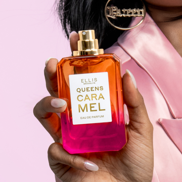Woman with black hair wearing a pink satin blazer and gold queen hoops holds QUEENS CARAMEL Eau de Parfum 50ml without cap on pink background