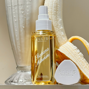 BANANA MILKSHAKE Perfume Mist 90ml without cap next to it's cap, a half peeled banana, and a vanilla milkshake in a clear glass cup on beige background