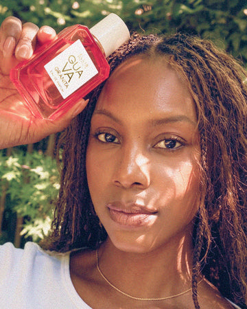 Woman with brown hair wearing a gold necklace and white t shirt holding GUAVA GRANITA Eau de Parfum 50ml next to her head against a background of green leaves