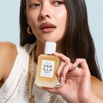 VANILLA SALT 50ml Eau de Parfum bottle held by a woman wearing a white lace top, pearl necklaces, and gold jewelry.