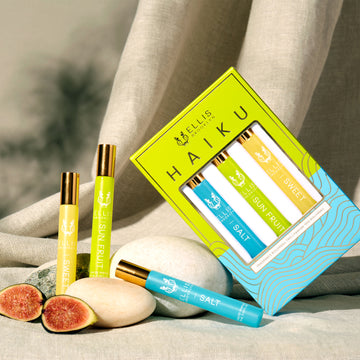 Boîte trio de roll-on parfum Haiku vert, bleu et or montrant le roll-on parfum Sel 10ml, le roll-on parfum Fruit du Soleil 10ml et le roll-on parfum Douceur 10ml, à côté du roll-on parfum Sel 10ml, du roll-on parfum Fruit du Soleil 10ml, du roll-on parfum Douceur 10ml, et de figues posées sur des pierres, sur un fond de tissu beige.