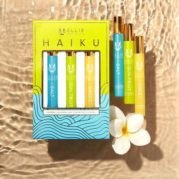 Green, blue, and gold Haiku Fragrance Rollerball Trio Box showing Salt 10ml Fragrance Rollerball, Sun Fruit 10ml Fragrance Rollerball, and Sweet 10ml Fragrance Rollerball, on beige water look backdrop with Salt 10ml Fragrance Rollerball, Sun Fruit 10ml Fragrance Rollerball, Sweet 10ml Fragrance Rollerball, and white flower.