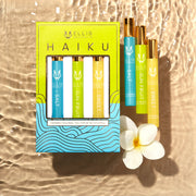 Boîte trio de flacons roll-on parfumés Haiku vert, bleu et or, comprenant le roll-on parfum Salt 10ml, le roll-on parfum Sun Fruit 10ml et le roll-on parfum Sweet 10ml, sur un fond beige effet eau avec les roll-ons Salt 10ml, Sun Fruit 10ml, Sweet 10ml et une fleur blanche.