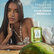 Model with brown hair wearing a white shell top and white skirt lays on her stomach in wet sand with her hand on her chin behind ISLA SIRENA Eau de Parfum 50ml sitting on top of a green coconut with white text that reads 