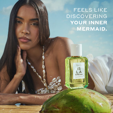 Model with brown hair wearing a white shell top and white skirt lays on her stomach in wet sand with her hand on her chin behind ISLA SIRENA Eau de Parfum 50ml sitting on top of a green coconut with white text that reads 