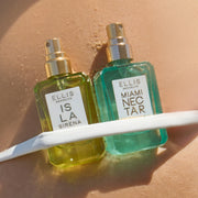 Two eau de parfum bottles, ISLA SIRENA 50mL & MIAMI NECTAR 50mL tucked between a white bikini bottom strap with caps off showcasing gold spray.