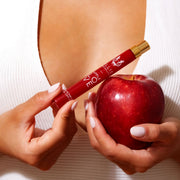 Model with light pink nail polish wearing white shirt holding Apple Love 10ml Eau De Parfum Travel Spray in one hand and an apple in the other hand, near chest.