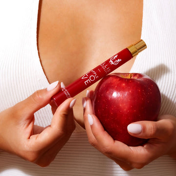 Mannequin avec du vernis à ongles rose clair portant une chemise blanche, tenant le spray de voyage Apple Love 10ml Eau De Parfum dans une main et une pomme dans l'autre main, près de la poitrine.