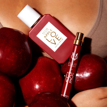 Model wearing brown tank top holding apples, Apple Love 50ml Eau De Parfum, and Apple Love 10ml Eau De Parfum Travel Spray against chest.