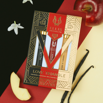 Boîte trio de roll-on parfum Love Triangle en noir, rouge, beige et or, présentant le roll-on parfum Myth 10 ml, le roll-on parfum Apple Love 10 ml et le roll-on parfum Vanilla Milk 10 ml, sur fond noir, rouge et beige avec des fleurs blanches, une pomme et des gousses de vanille.