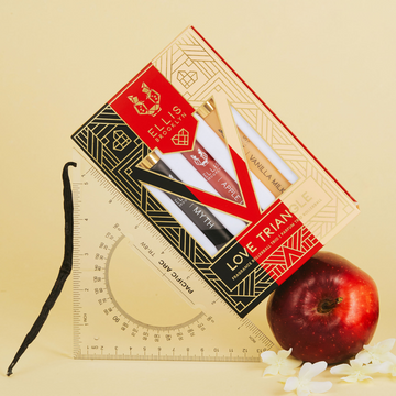 Boîte trio de roll-on parfum Love Triangle en noir, rouge, beige et or, montrant le roll-on parfum Myth 10 ml, le roll-on parfum Apple Love 10 ml et le roll-on parfum Vanilla Milk 10 ml, appuyés sur un outil de mesure et une pomme avec une gousse de vanille et des fleurs blanches, sur un fond jaune clair.