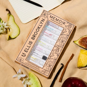 Memoir Fragrance Discovery Set box with beige and black floral design showing Dear Sky 1.5ml Vial, Miami Nectar 1.5ml Vial, Apple Love 1.5ml Vial, Salt 1.5ml Vial, Sun Fruit 1.5ml Vial, Florist 1.5ml Vial, Myth 1.5ml Vial, Sweet 1.5ml Vial, and Vanilla Milk 1.5ml Vial, on tan fabric with fig, pineapple, apple, sandalwood, vanilla bean, pear, white flowers, and notebook with pen.
