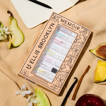 Memoir Fragrance Discovery Set box with beige and black floral design showing Dear Sky 1.5ml Vial, Miami Nectar 1.5ml Vial, Apple Love 1.5ml Vial, Salt 1.5ml Vial, Sun Fruit 1.5ml Vial, Florist 1.5ml Vial, Myth 1.5ml Vial, Sweet 1.5ml Vial, and Vanilla Milk 1.5ml Vial, on tan fabric with fig, pineapple, apple, sandalwood, vanilla bean, pear, white flowers, and notebook with pen.