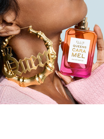 QUEENS CARAMEL 50ml Eau de Parfum bottle held by a woman wearing a pink sweater and oversized gold hoop earrings that say "QUEEN".